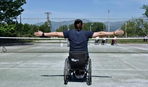 Cover. Tennis court. Back of a male wheelchair user stretching his arms left and right on the same line with the horizon.