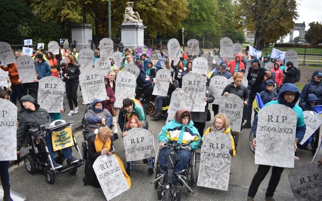 photo in exterior space, brusells park, where dozens of disabled or able-bodied people look at the camera holding paper signs that look like tumbstones and write R. I. P. 27.09.2022
